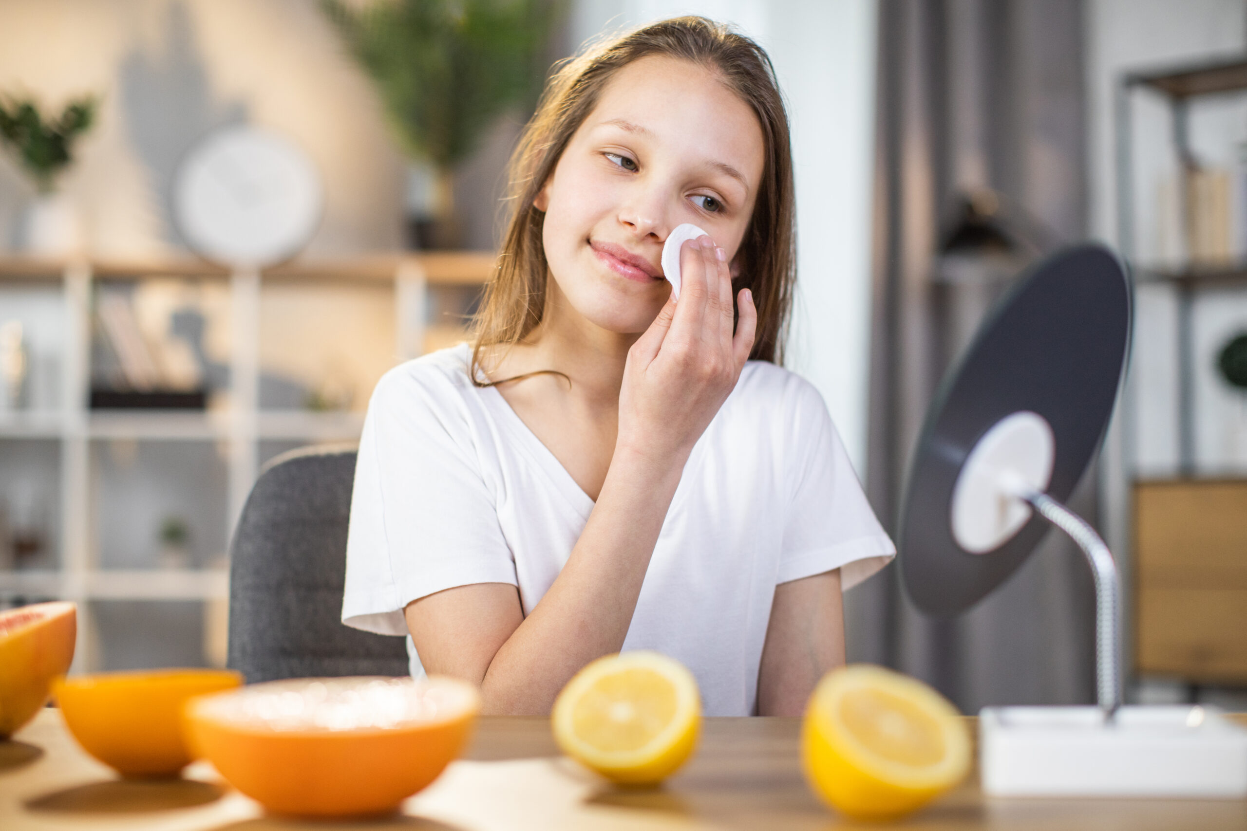Pretty female teenger sitting at table while cleaning face with cotton pad and looking at mirror. Happy young woman taking care of her healthy pure skin at home.