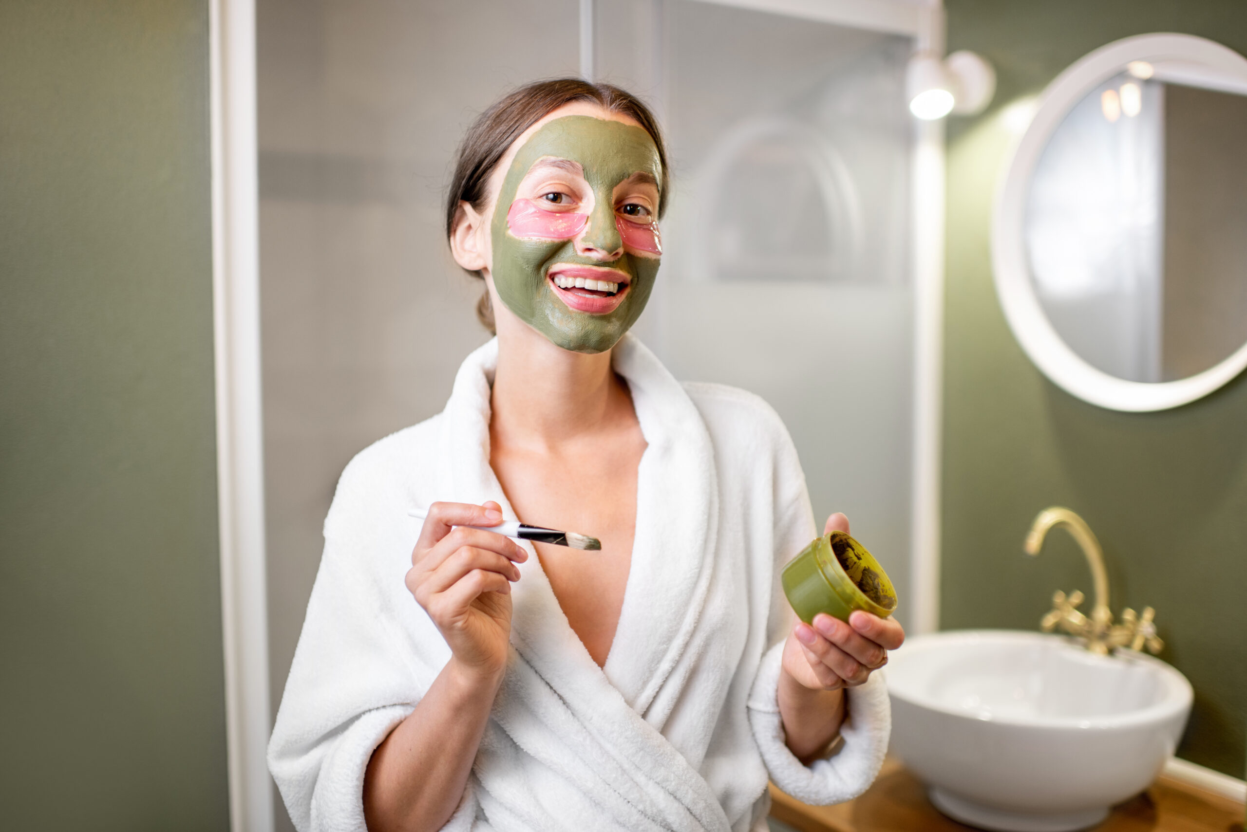 Beautiful woman taking care of herself with facial mask and patches under her eyes, standing in bathrobe in the modern bathroom
